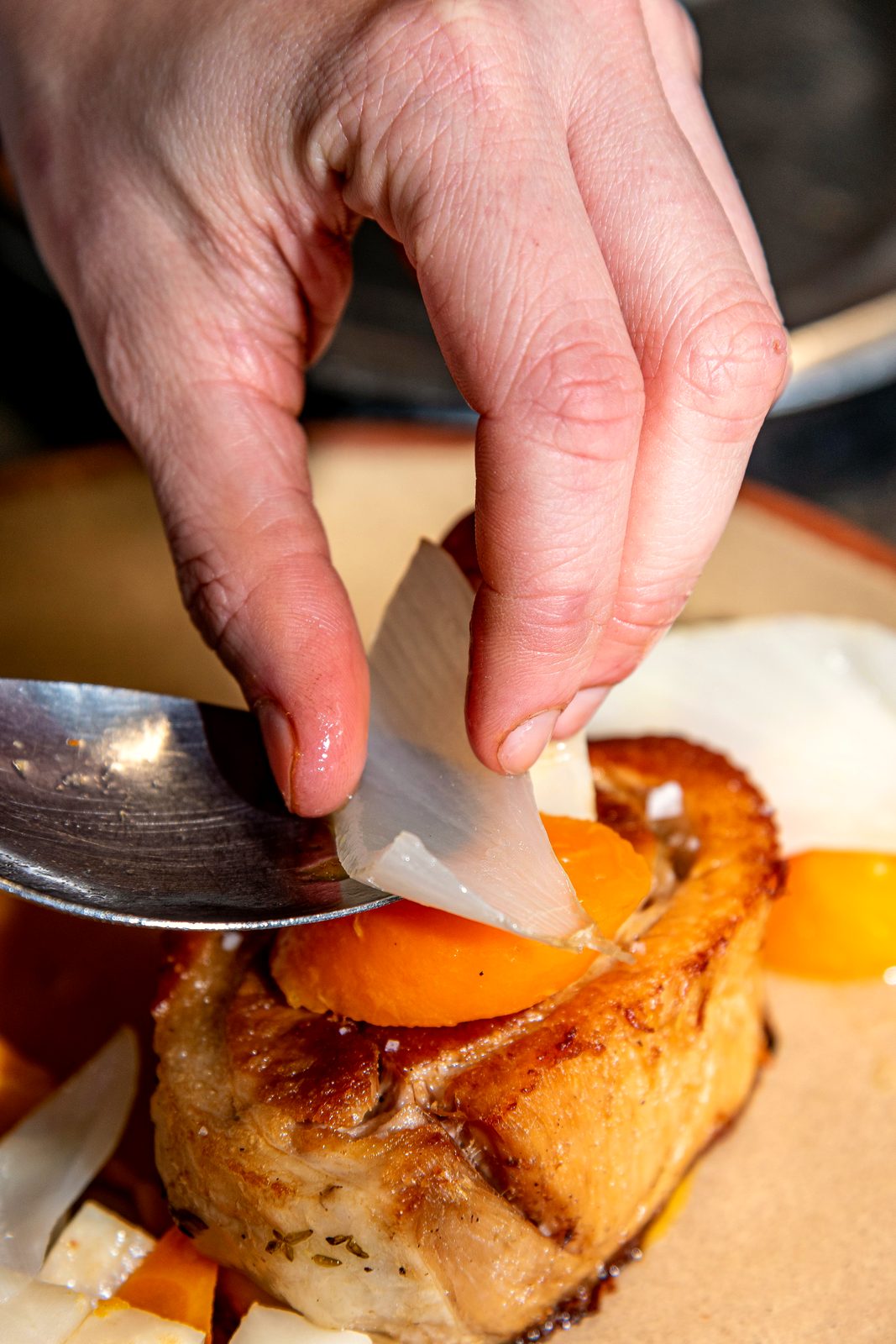 Les mains du chef déposent une tuile croustillante sur un plat au dressage, cuillère en appui, dans la cuisine du restaurant L’Apibo à Paris