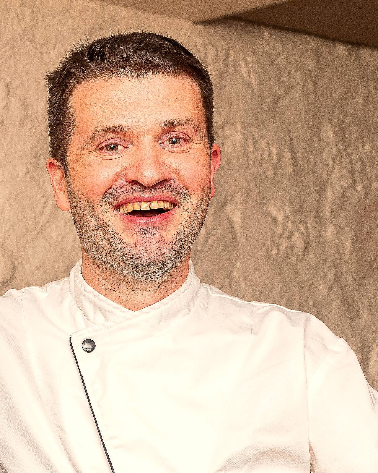 Portrait d’Anthony Boucher, chef et propriétaire de L’Apibo, en veste de cuisine blanche, souriant devant le mur de pierre claire de la salle, rue Tiquetonne à Paris 2e