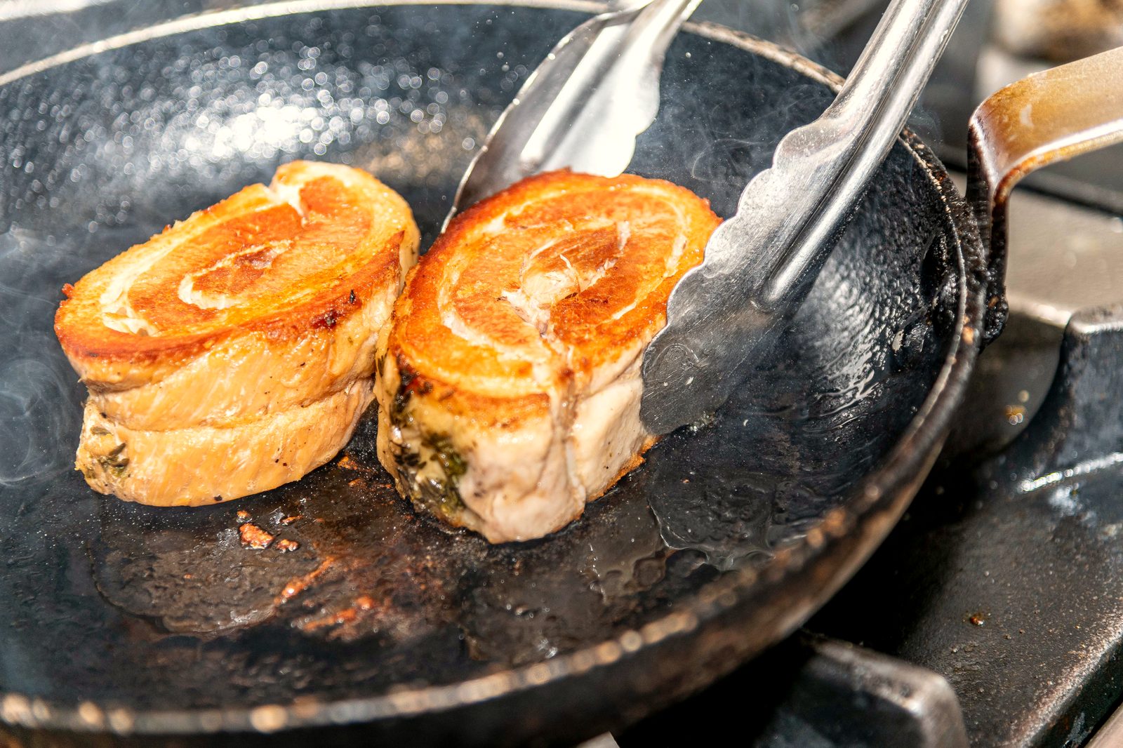 Deux pièces de cochon dorées saisies à la poêle dans la cuisine de L’Apibo, vapeur montante et pinces en main pendant le service, à Paris