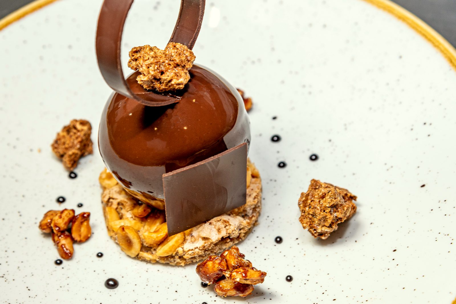 Dessert au chocolat de L’Apibo : sphère brillante, ruban de chocolat, noix caramélisées et pointes de ganache sur assiette mouchetée, à Paris