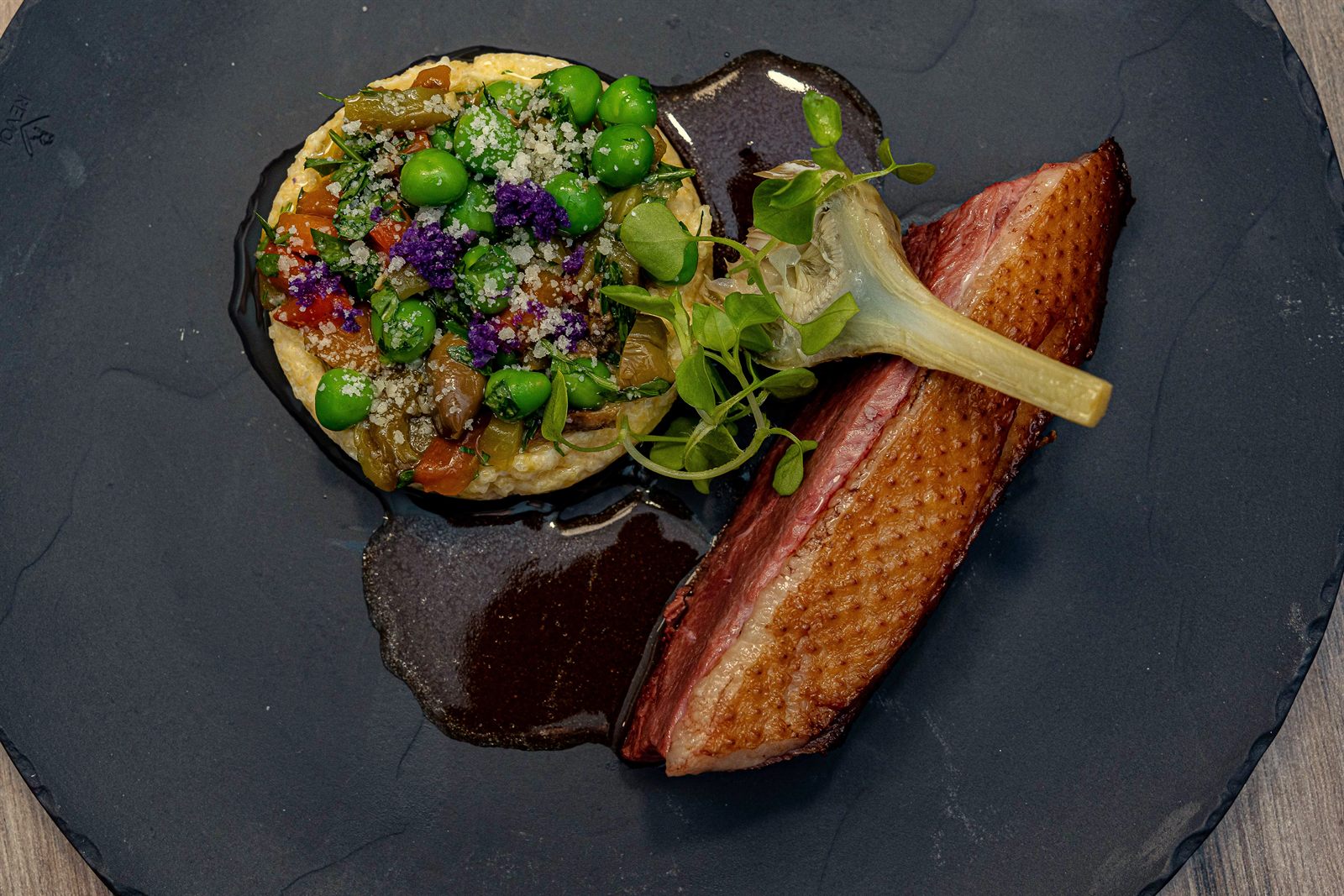 Magret de canard rôti à la peau croustillante, petits pois, cœur d’artichaut et jus corsé, dressé sur ardoise sombre au restaurant L’Apibo à Paris 2e