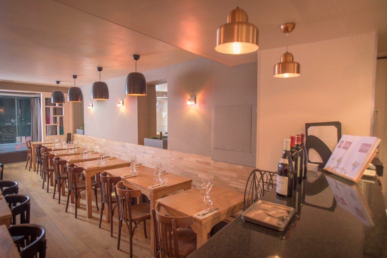 Salle du restaurant L’Apibo rue Tiquetonne à Paris : murs de pierre blanchie, tables en bois clair dressées et suspensions noires diffusant une lumière chaude