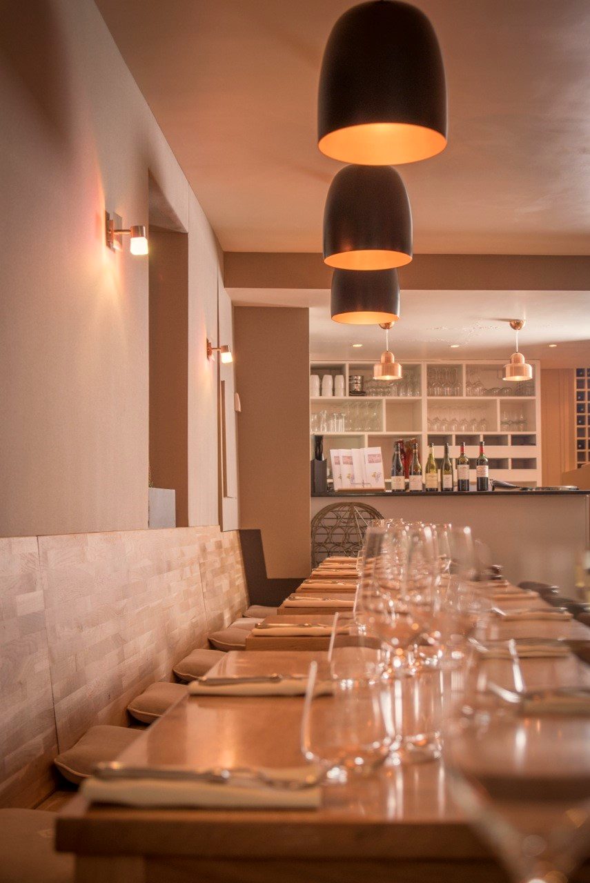 Perspective de la salle de L’Apibo vers le bar : tables en bois dressées, suspensions noires et vaisselle prête pour le service, à Paris 2e