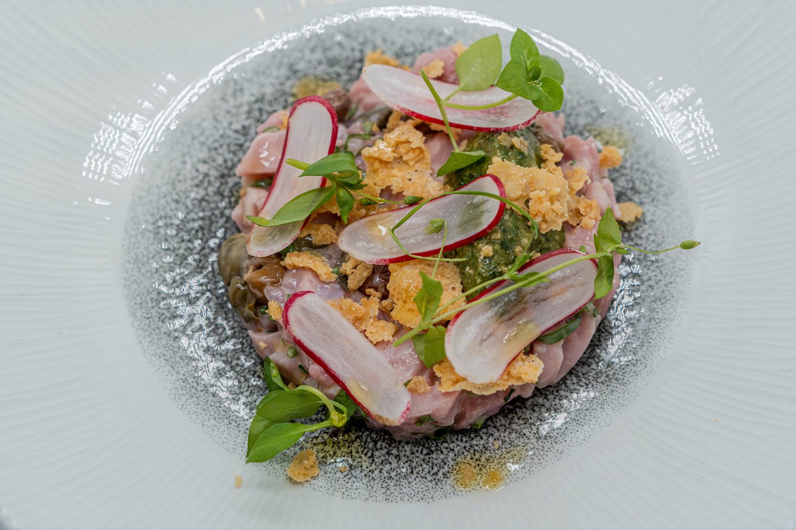 Tartare rosé relevé aux herbes, fines lamelles de radis rose, tuile croustillante dorée et jeunes pousses, dressé en dôme sur assiette gris moucheté au restaurant L’Apibo à Paris